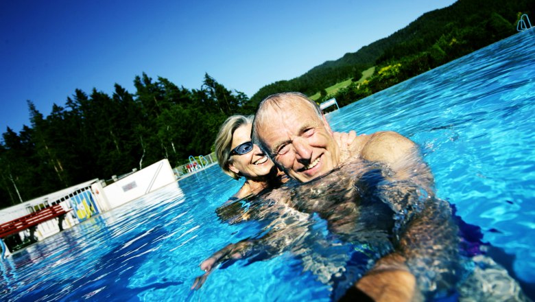 Mitterbach outdoor pool, © weinfranz.at An elderly couple smiling as they swim in an outdoor pool with a forest in the background.