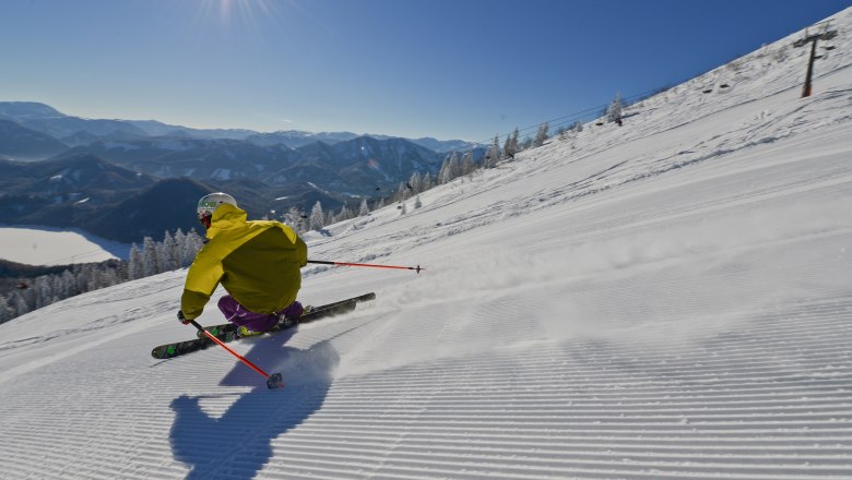 Gipfelhang mit Blick auf den Erlaufsee, © Bergbahnen Mitterbach Skifahrer auf einer Piste mit Blick auf den Erlaufsee und Berge im Hintergrund.