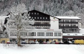 Ein verschneites Hotelgebäude mit der Aufschrift 'Sporthotel' und 'Cafeteria', umgeben von schneebedeckten Bäumen und geparkten Autos.