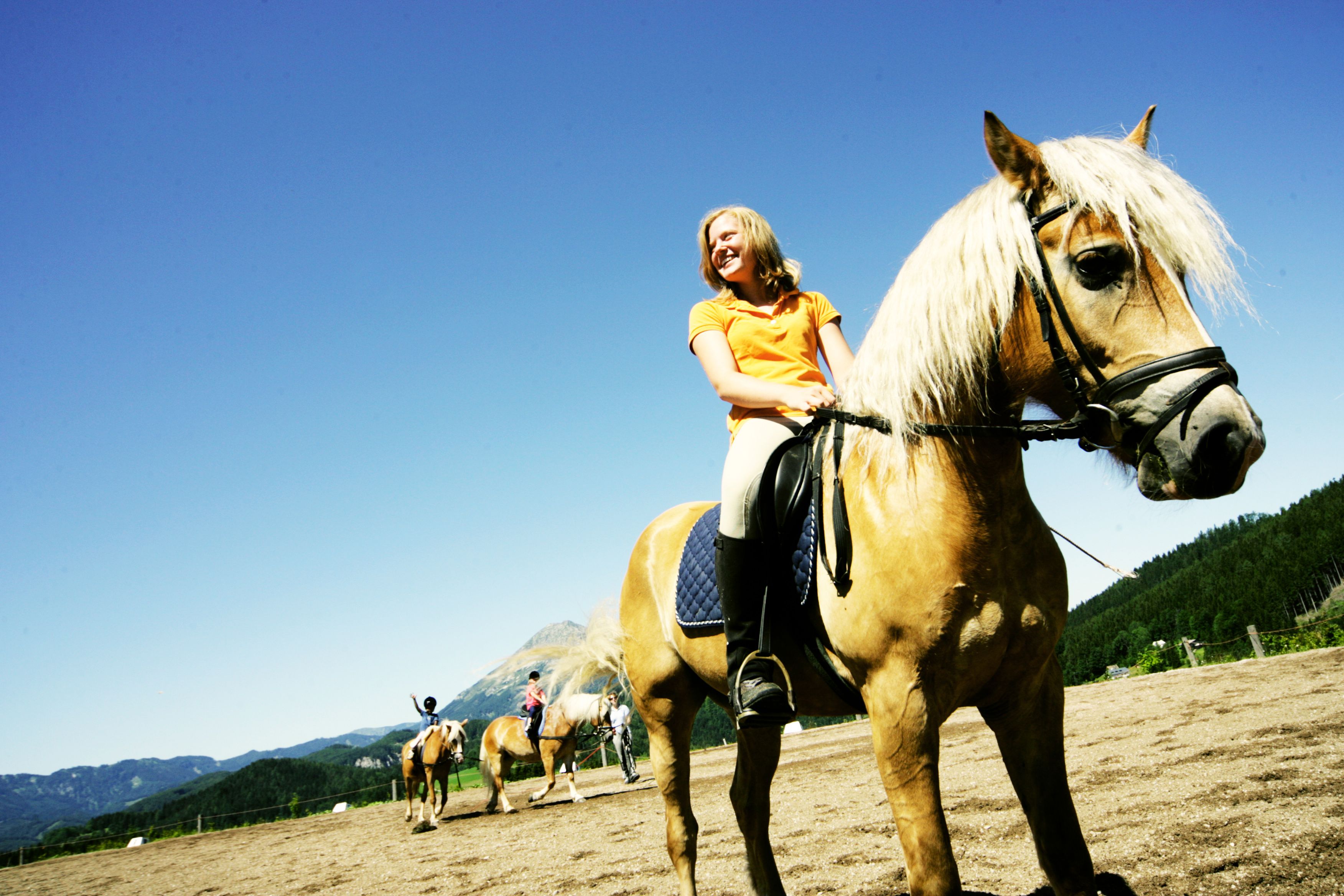 Eine Frau reitet auf einem braunen Pferd mit blonder Mähne auf einem Reitplatz, umgeben von Bergen und blauem Himmel.