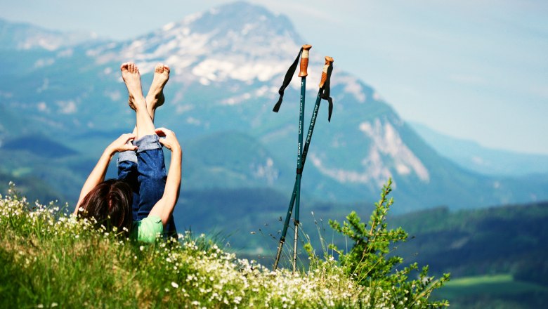 Person liegt auf einer Wiese mit Blick auf die Berge, Wanderst&ouml;cke daneben.