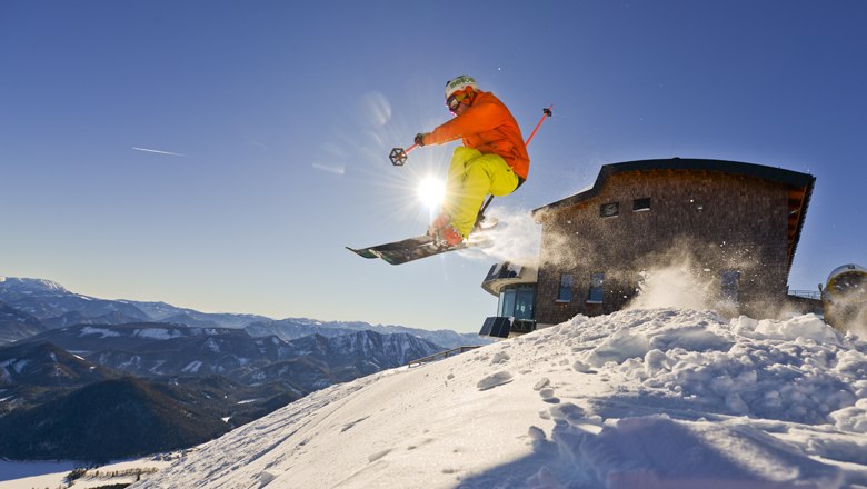 Action & Fun auf der Gemeindealpe-Terzerhaus, © Leiminger Ein Skifahrer springt vor dem Terzerhaus auf der Gemeindealpe in die Luft, umgeben von schneebedeckten Bergen und blauem Himmel.