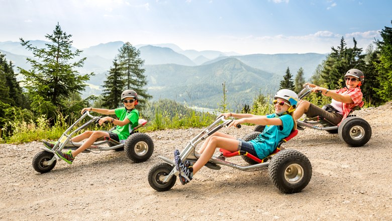 Three people ride down a mountain on mountain carts, surrounded by a mountain backdrop.
