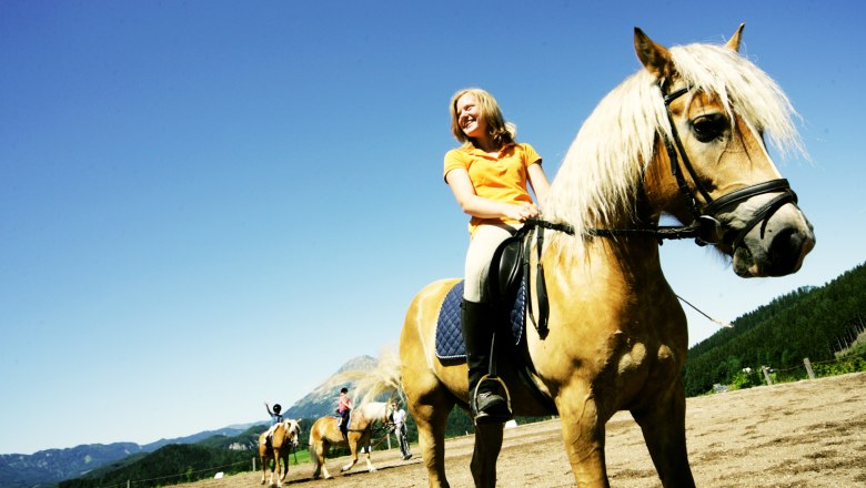 Eine Frau reitet auf einem braunen Pferd mit blonder Mähne auf einem Reitplatz, umgeben von Bergen und blauem Himmel.