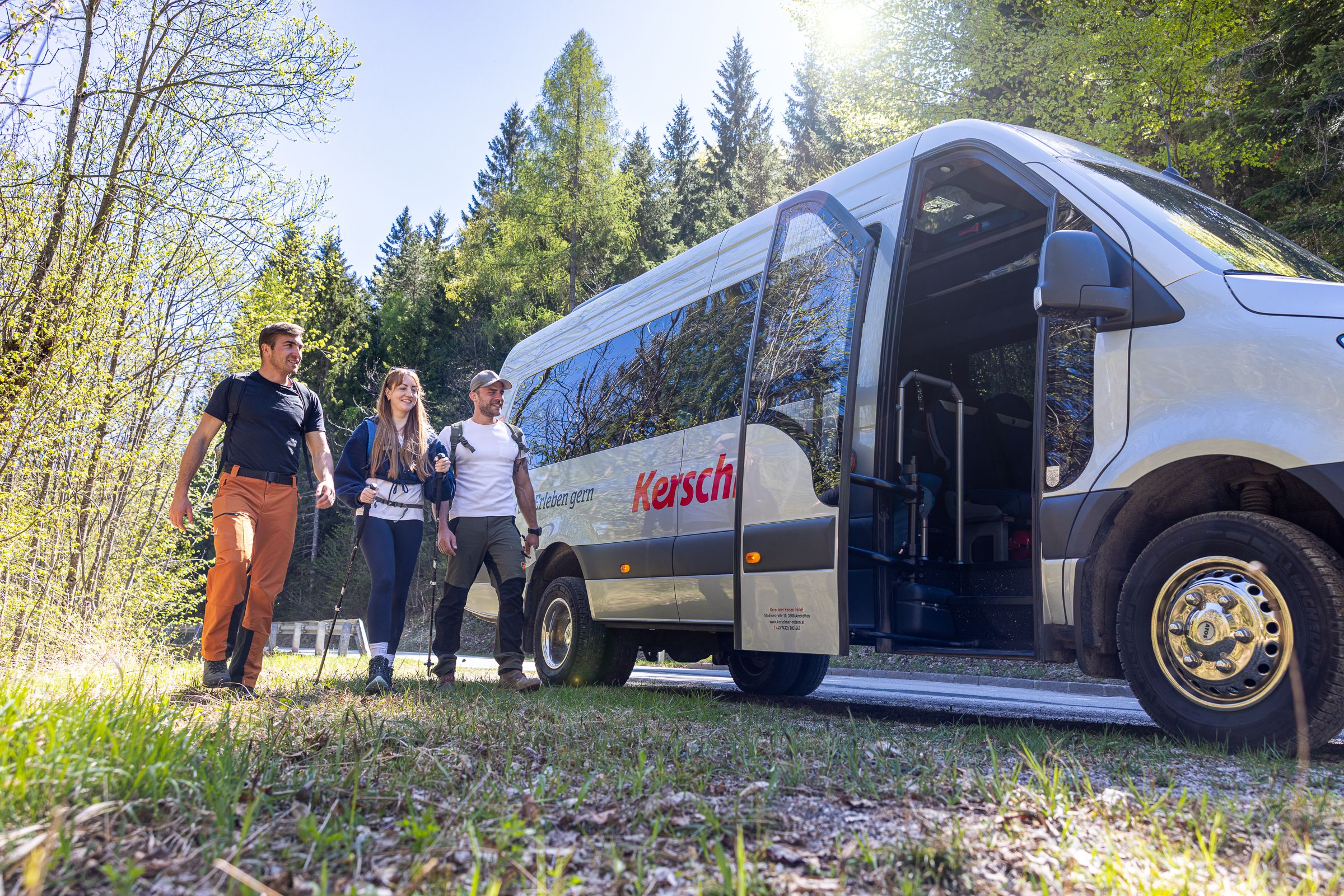 Drei wandernde Personen nähern sich einem Kleinbus, welcher bereits die Tür geöffnet hat. 