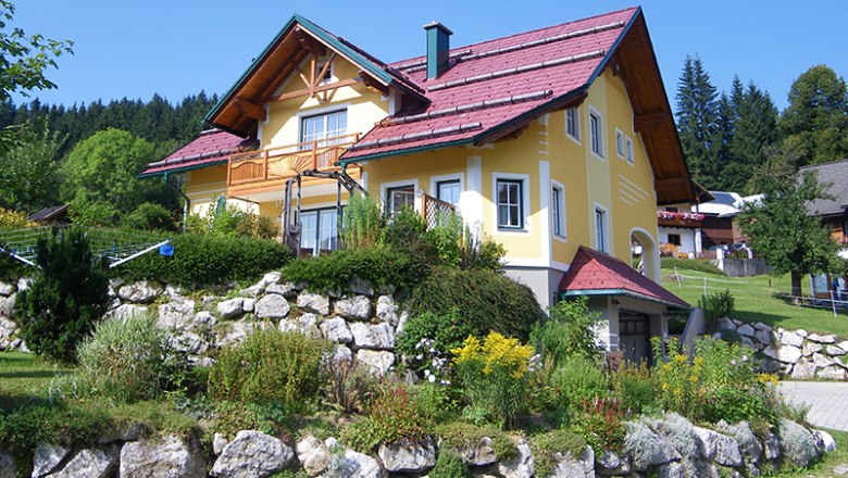 Gelbes Haus mit rotem Dach in einer grünen Landschaft.