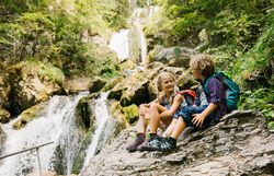 Zwei Kinder sitzen entspannt auf einem großen Stein und genießen die erfrischende Atmosphäre des Naturparks. Umgeben von üppigem Grün und dem sanften Rauschen des Wassers, strahlt die Szene pure Lebensfreude und Abenteuerlust aus.