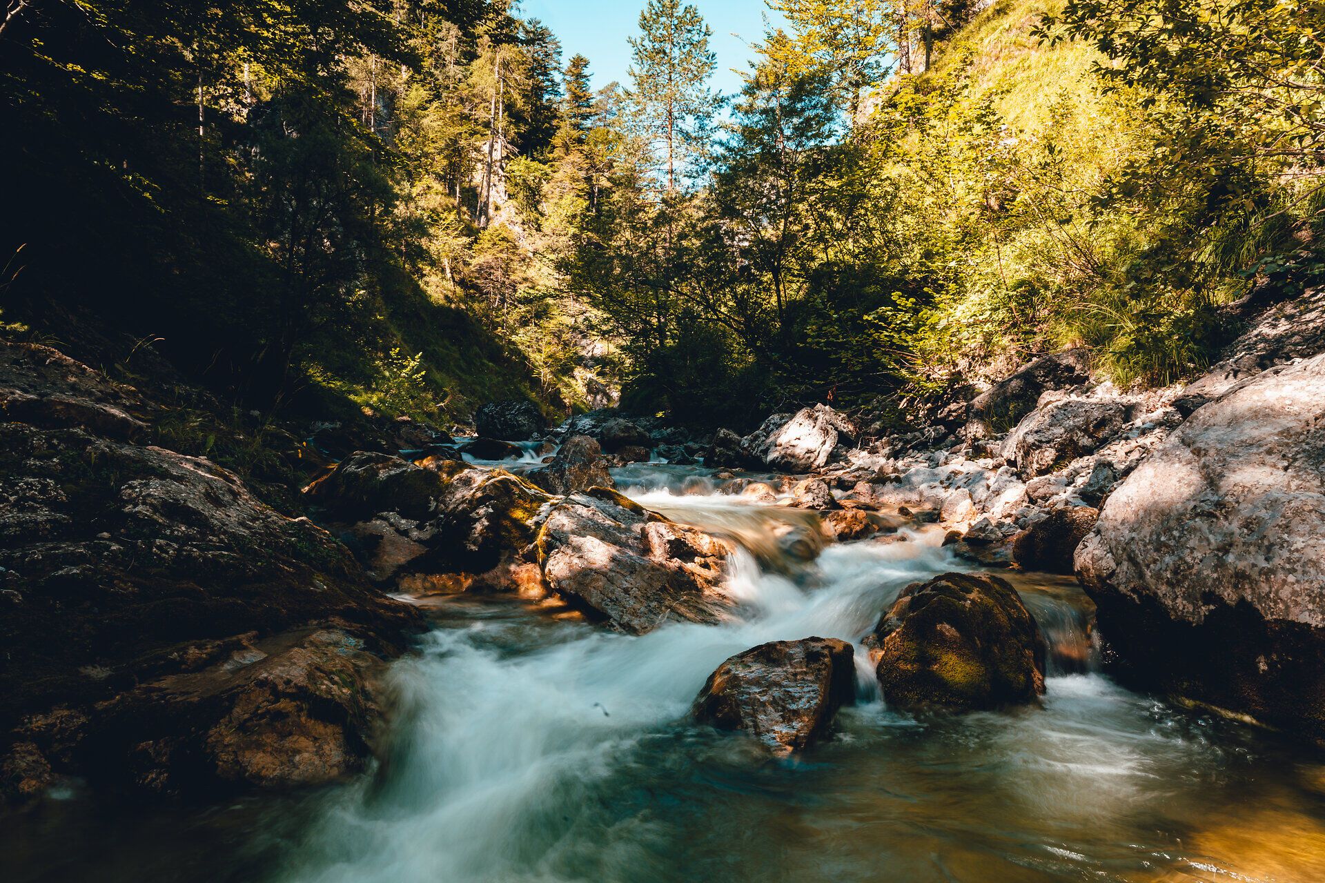 Ein sanft plätschernder Bach schlängelt sich durch die üppige, grüne Landschaft, umgeben von majestätischen Bäumen und schroffen Felsen. Die klare, frische Luft und das beruhigende Geräusch des Wassers laden zu einem entspannenden Spaziergang ein, während die Sonnenstrahlen durch das Blätterdach tanzen.