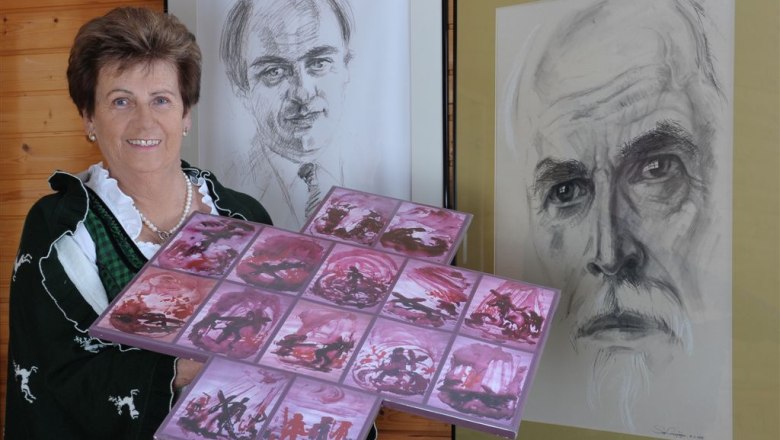 Frau in Tracht hält Kunstwerk vor zwei Porträts.