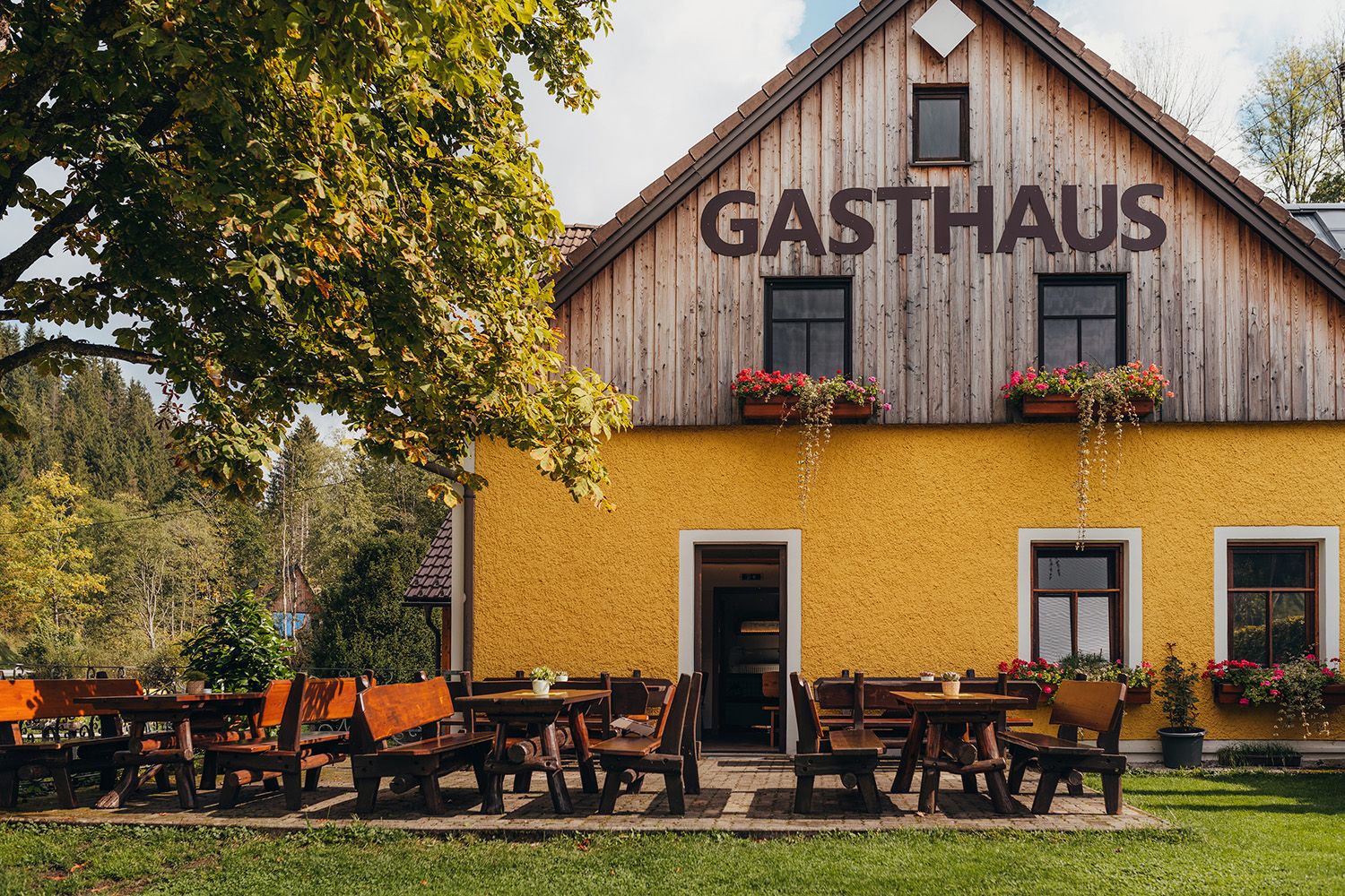Ein gelbes Gasthaus mit Holztischen und Bänken im Freien, umgeben von Bäumen und Natur.