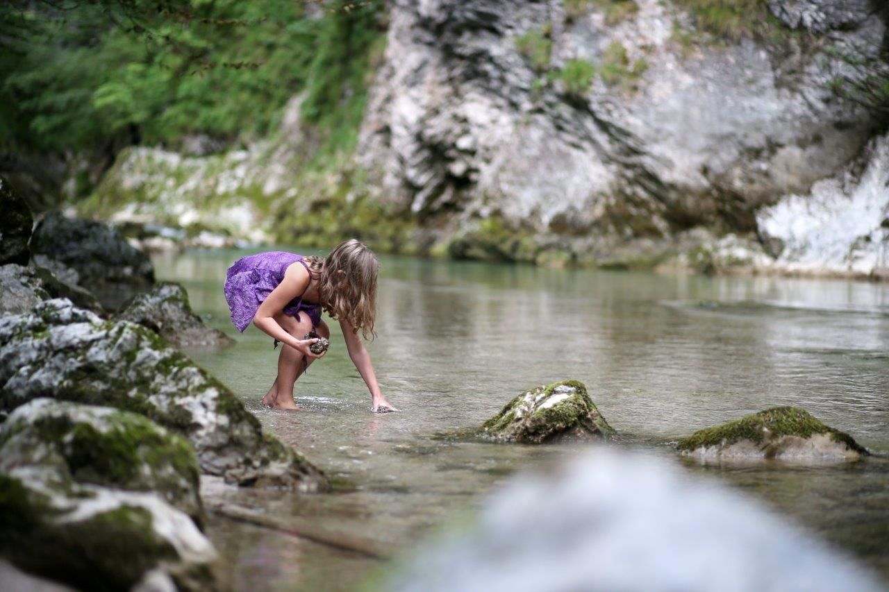 A child in a purple dress plays in a clear stream, surrounded by rocks and green nature.