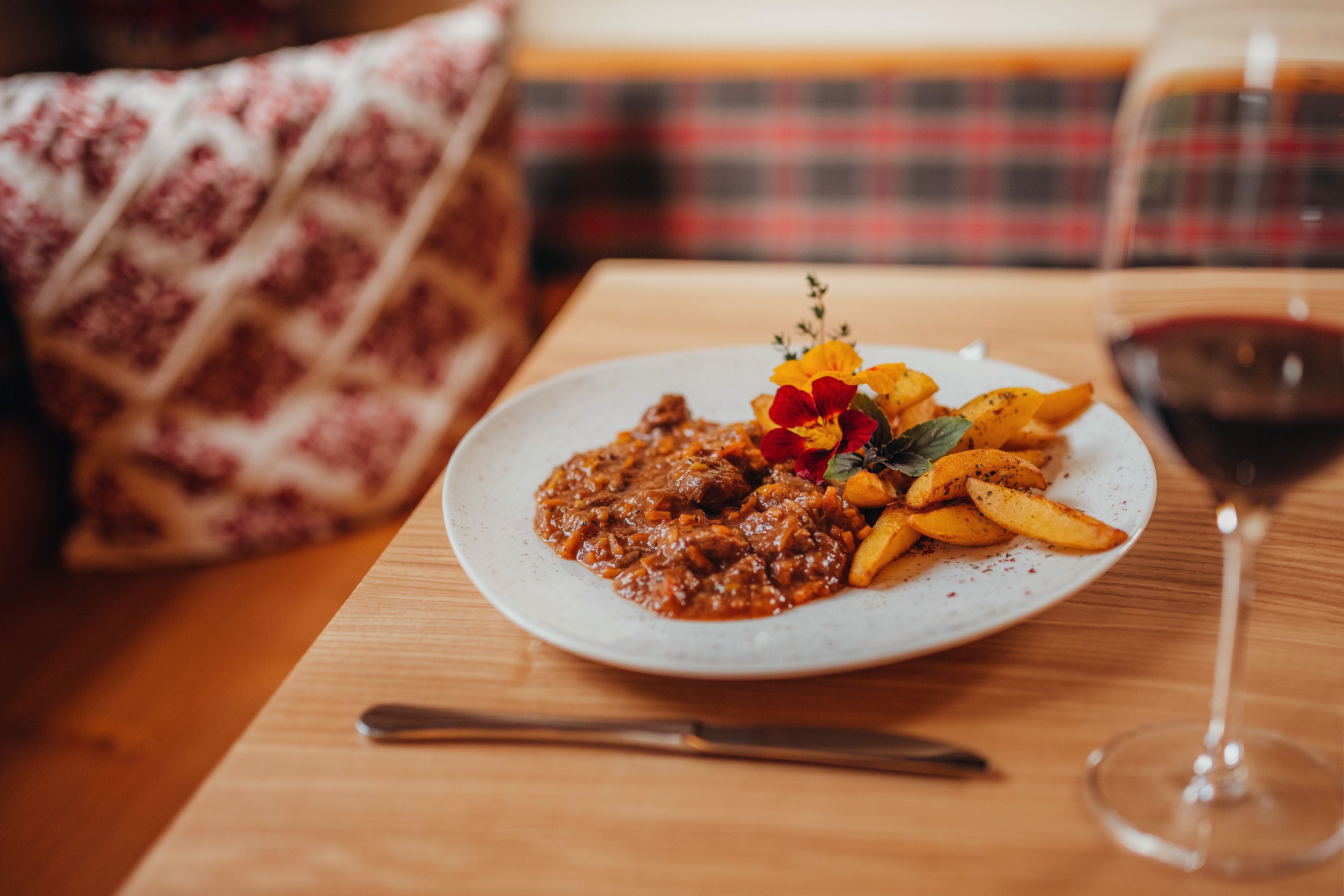 Ein Teller mit Lammragout und Thymian-Kartoffeln, dekoriert mit essbaren Blüten, auf einem Holztisch. Ein Glas Rotwein steht daneben.