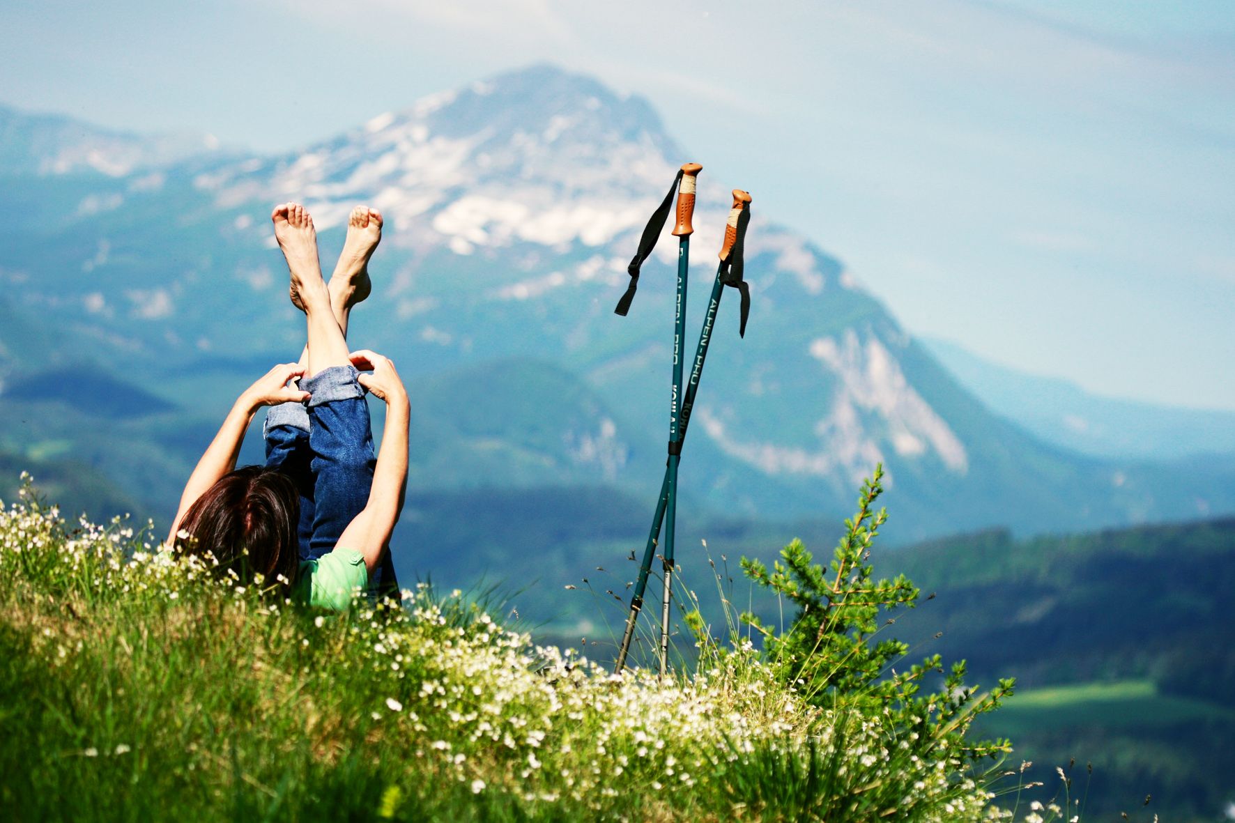Person lying on a meadow with a view of the mountains, hiking poles next to them.