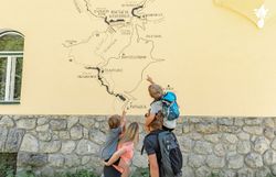 Eine Gruppe von Wanderern steht fasziniert vor einer großen Wandkarte, die die atemberaubenden Wanderwege des Naturparks zeigt. Die Kinder zeigen begeistert auf die verschiedenen Routen, während die Sonne sanft auf die gelbe Wand scheint und die Vorfreude auf das Abenteuer in der Natur spürbar ist.