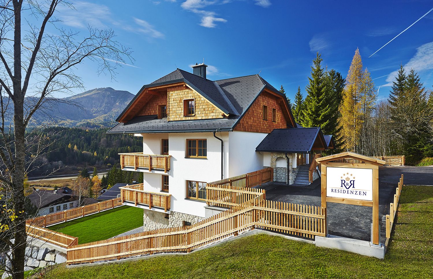 A modern house in an alpine setting with wooden cladding and balconies, surrounded by trees and mountains. A sign shows 'R&R Residences'.