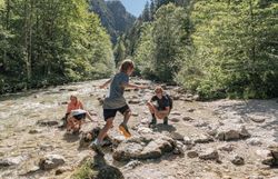 Die Sonne strahlt über den glitzernden Fluss, während fröhliche Kinder von Stein zu Stein hüpfen. Umgeben von üppigem Grün und majestätischen Bäumen, lädt diese idyllische Landschaft zum Verweilen und Entdecken ein. Hier wird der Sommer in den Bergen zum unvergesslichen Erlebnis.