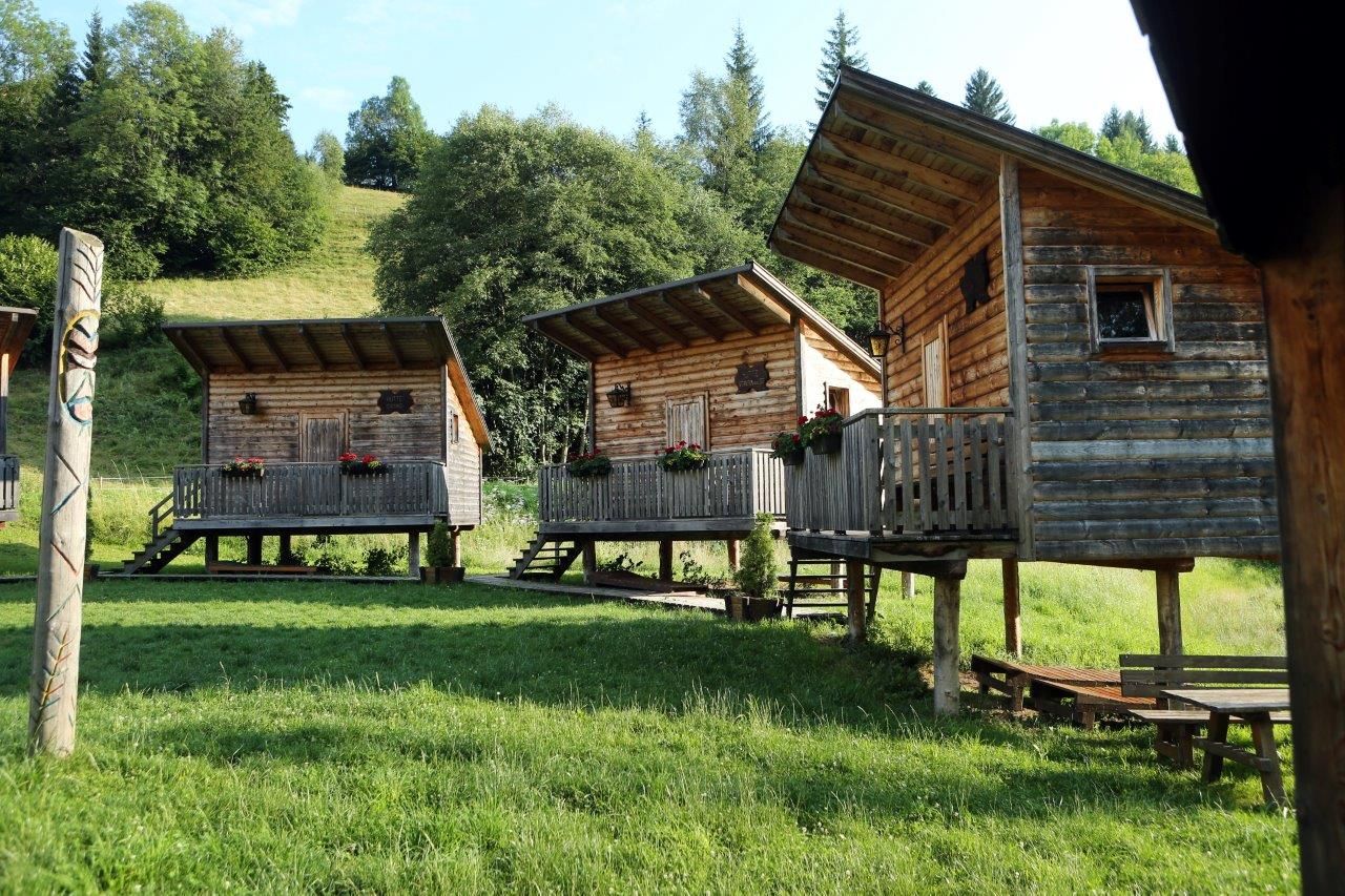 Drei Holzhütten auf Stelzen mit Veranda in einem grünen, bewaldeten Gebiet.