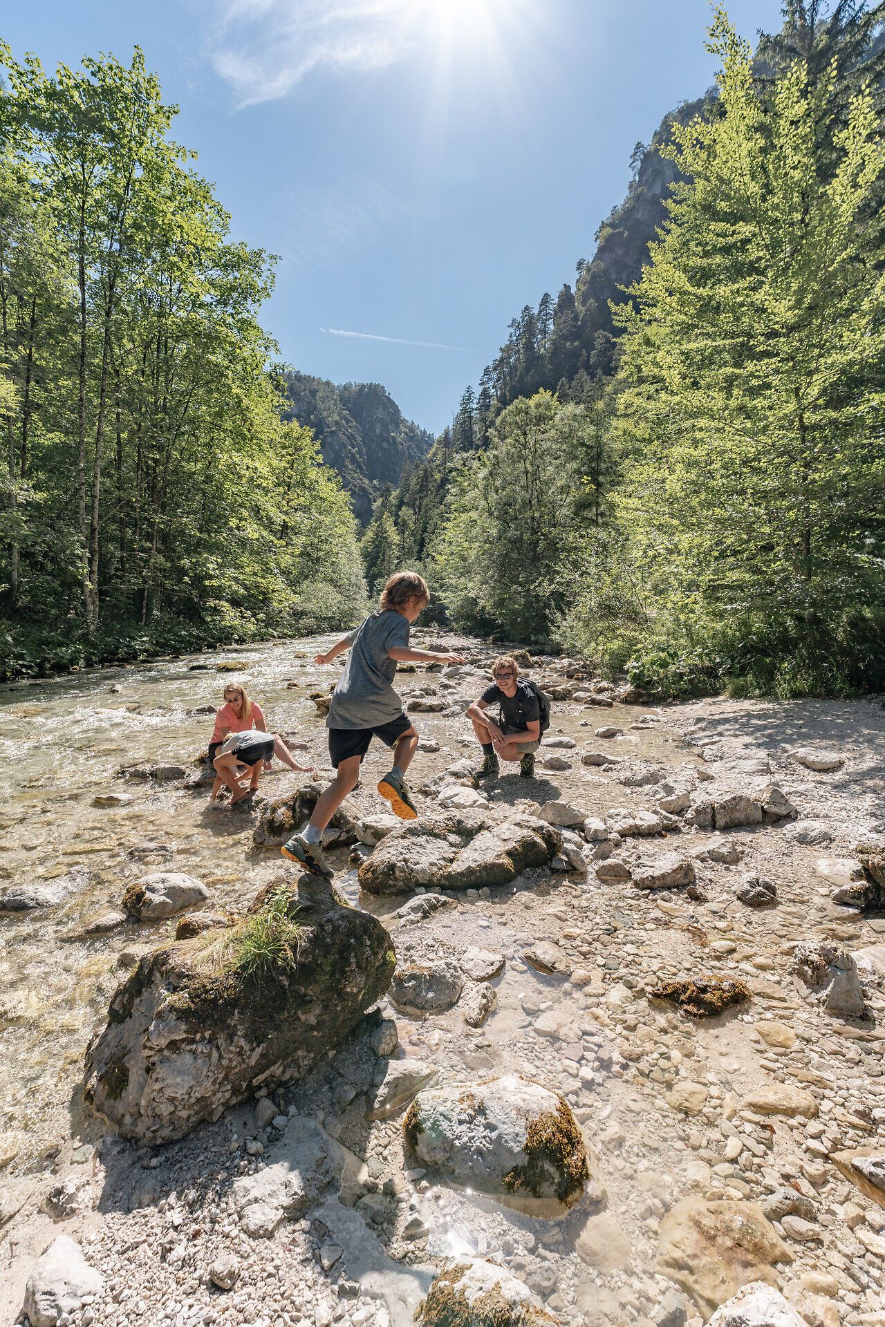 Die Sonne strahlt über den glitzernden Fluss, während fröhliche Kinder von Stein zu Stein hüpfen. Umgeben von üppigem Grün und majestätischen Bäumen, lädt diese idyllische Landschaft zum Verweilen und Entdecken ein. Hier wird der Sommer in den Bergen zum unvergesslichen Erlebnis.