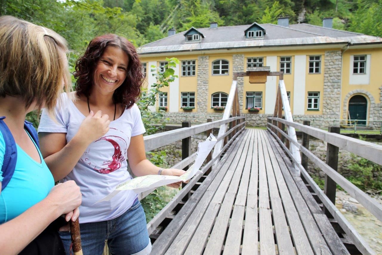 Zwei Frauen stehen auf einer Holzbrücke vor einem gelben Gebäude in Wienerbruck, eine hält eine Karte.