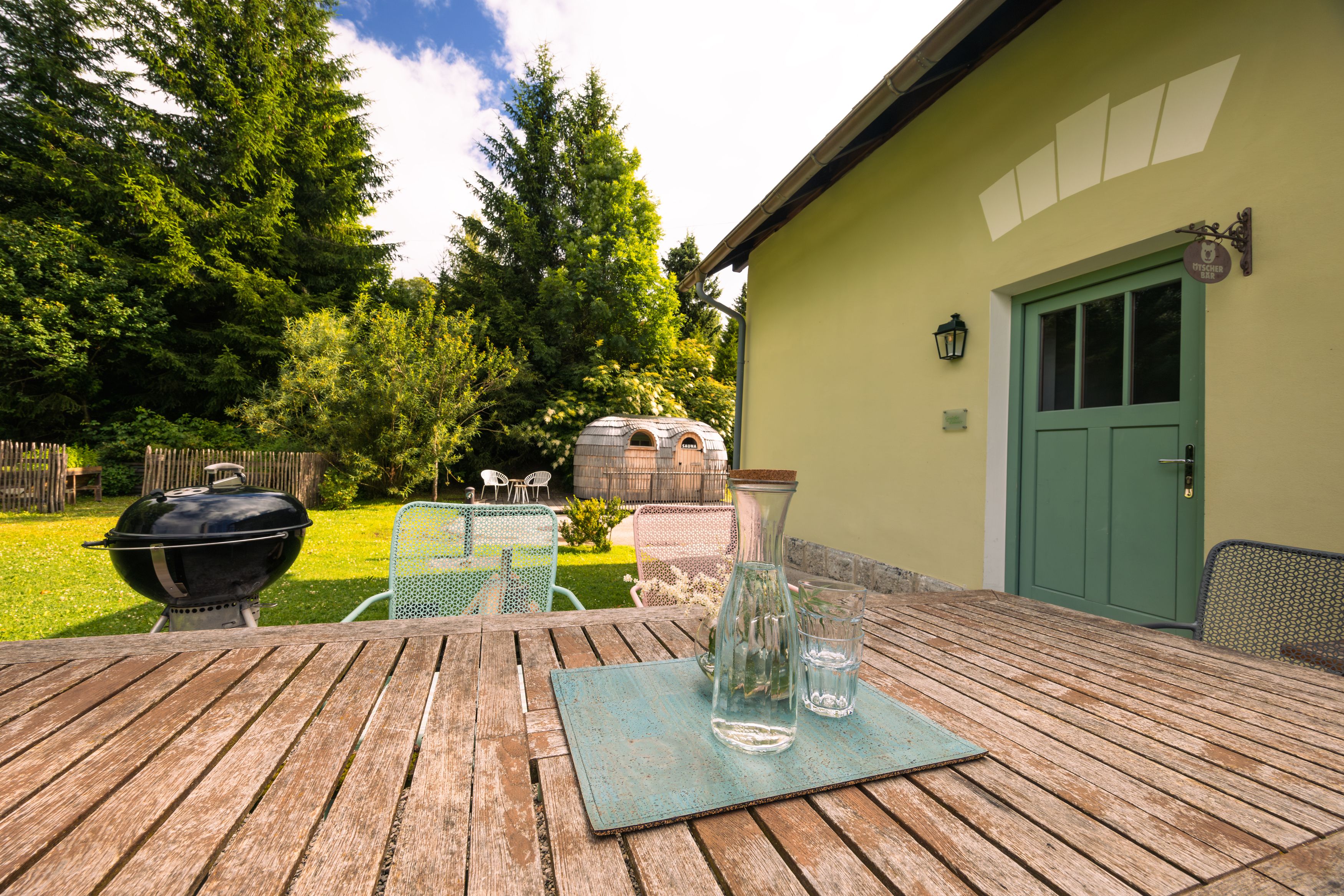 A garden with wooden table, chairs, barbecue and a barrel-shaped sauna.