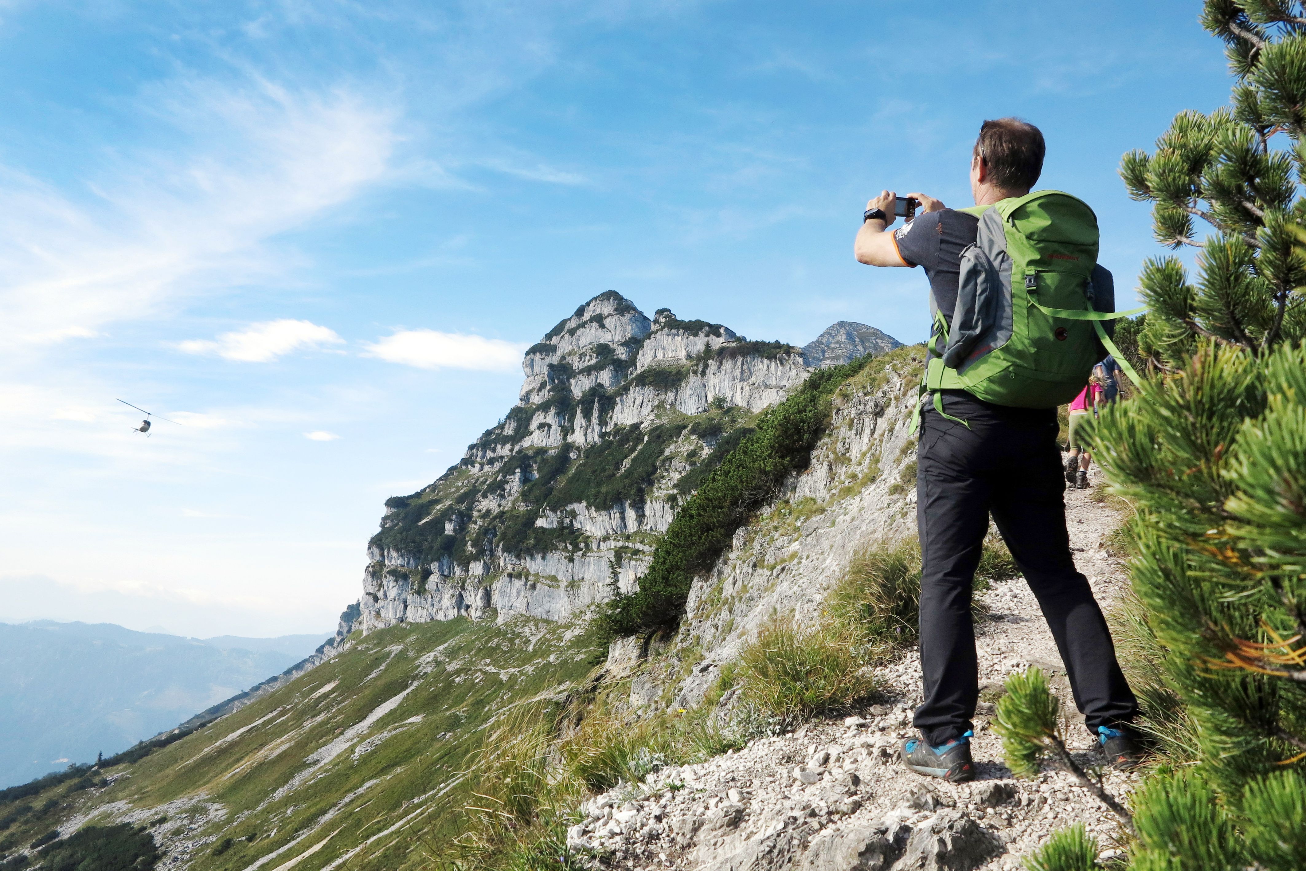 Ein Wanderer mit grünem Rucksack fotografiert eine Berglandschaft mit einem Hubschrauber am Himmel.