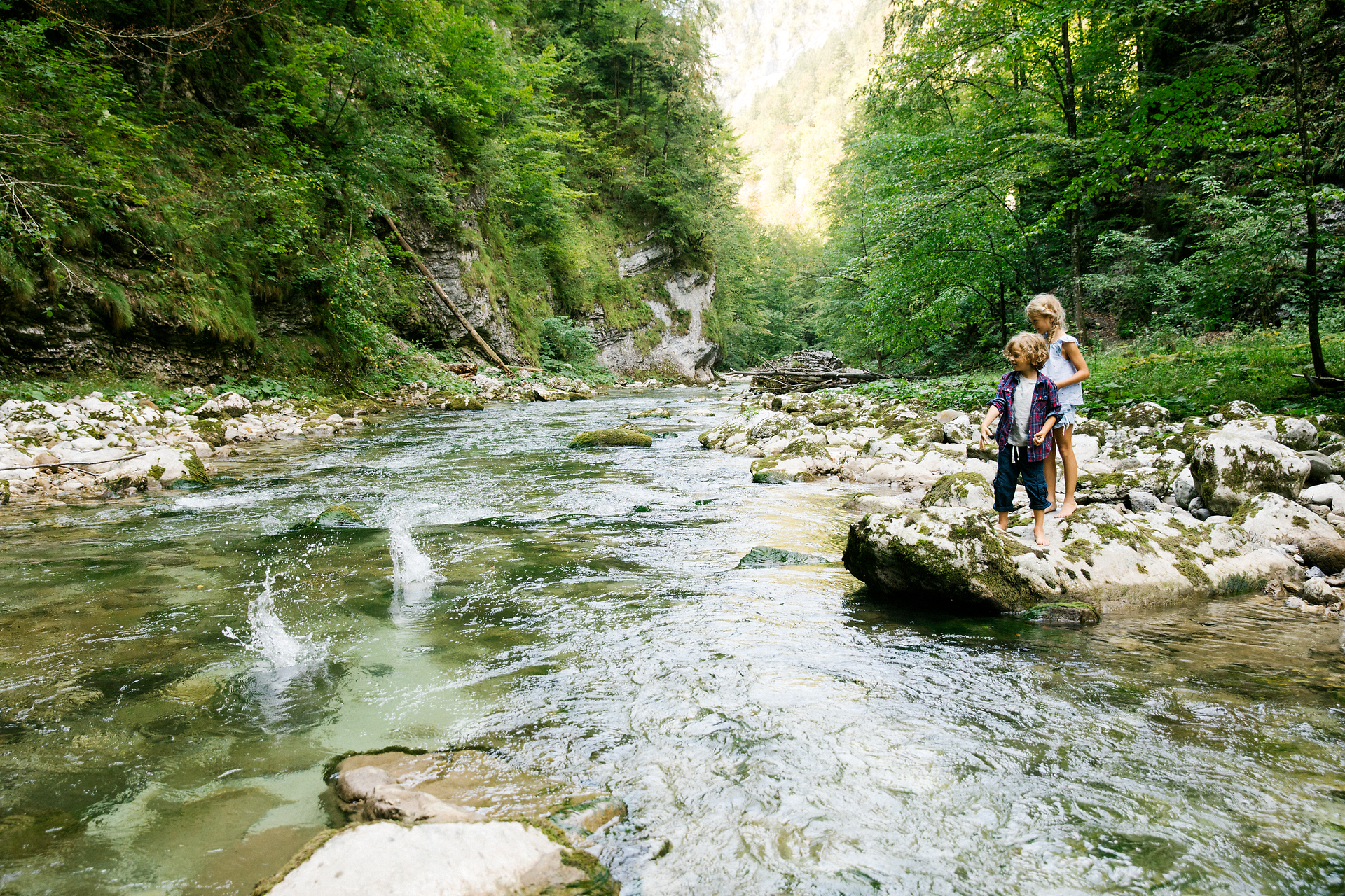 Ein erfrischender Fluss schlängelt sich durch die grüne Landschaft, während Kinder fröhlich am Ufer spielen. Die sanften Geräusche des Wassers und das Zwitschern der Vögel schaffen eine harmonische Atmosphäre, die zum Verweilen einlädt.