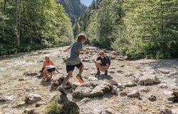 Die Sonne strahlt über den glitzernden Fluss, während fröhliche Kinder von Stein zu Stein hüpfen. Umgeben von üppigem Grün und majestätischen Bäumen, lädt diese idyllische Landschaft zum Verweilen und Entdecken ein. Hier wird der Sommer in den Bergen zum unvergesslichen Erlebnis.
