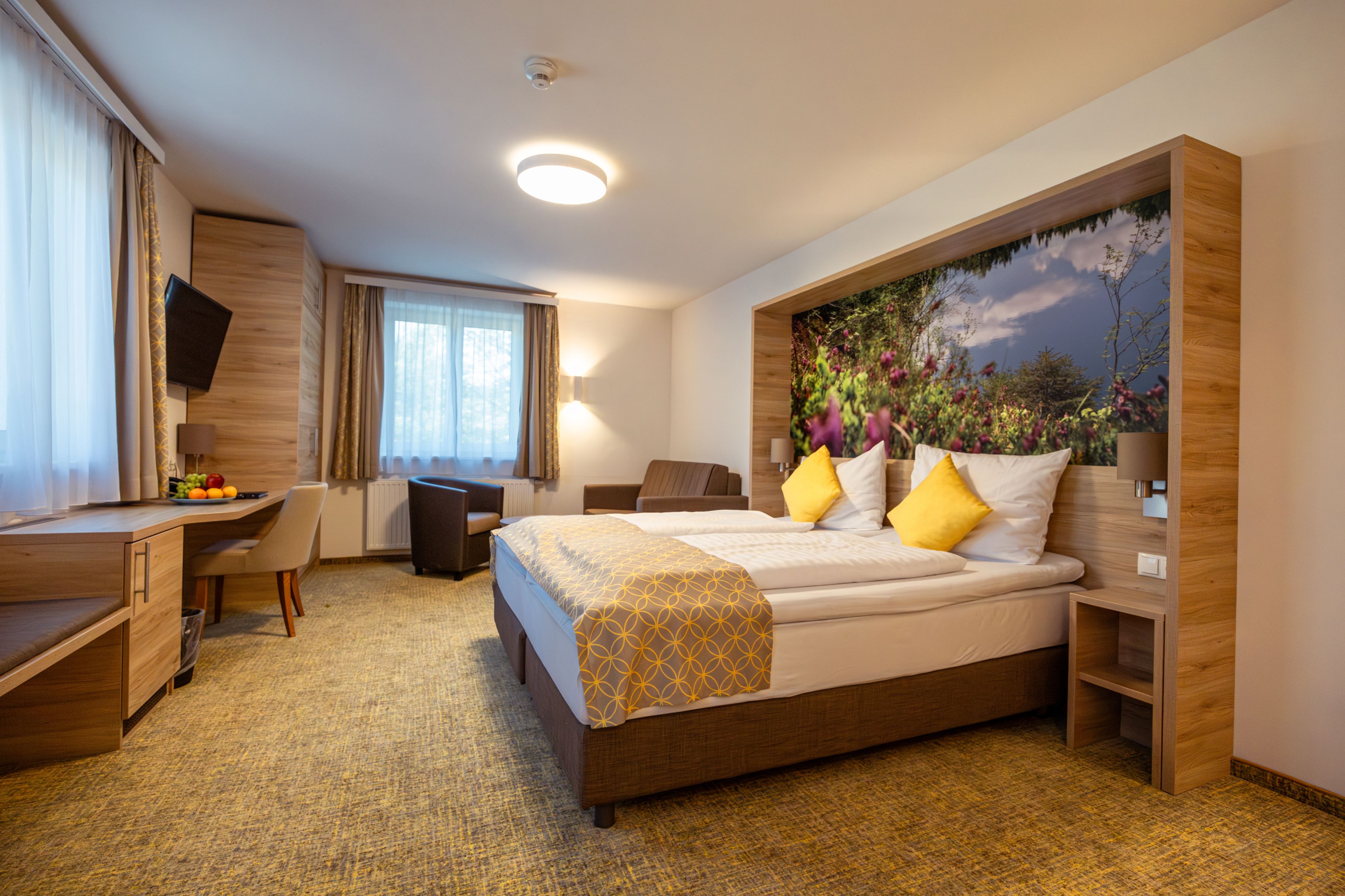 Modernes Hotelzimmer mit Doppelbett, gelben Kissen, Schreibtisch, Sessel und großem Fenster.