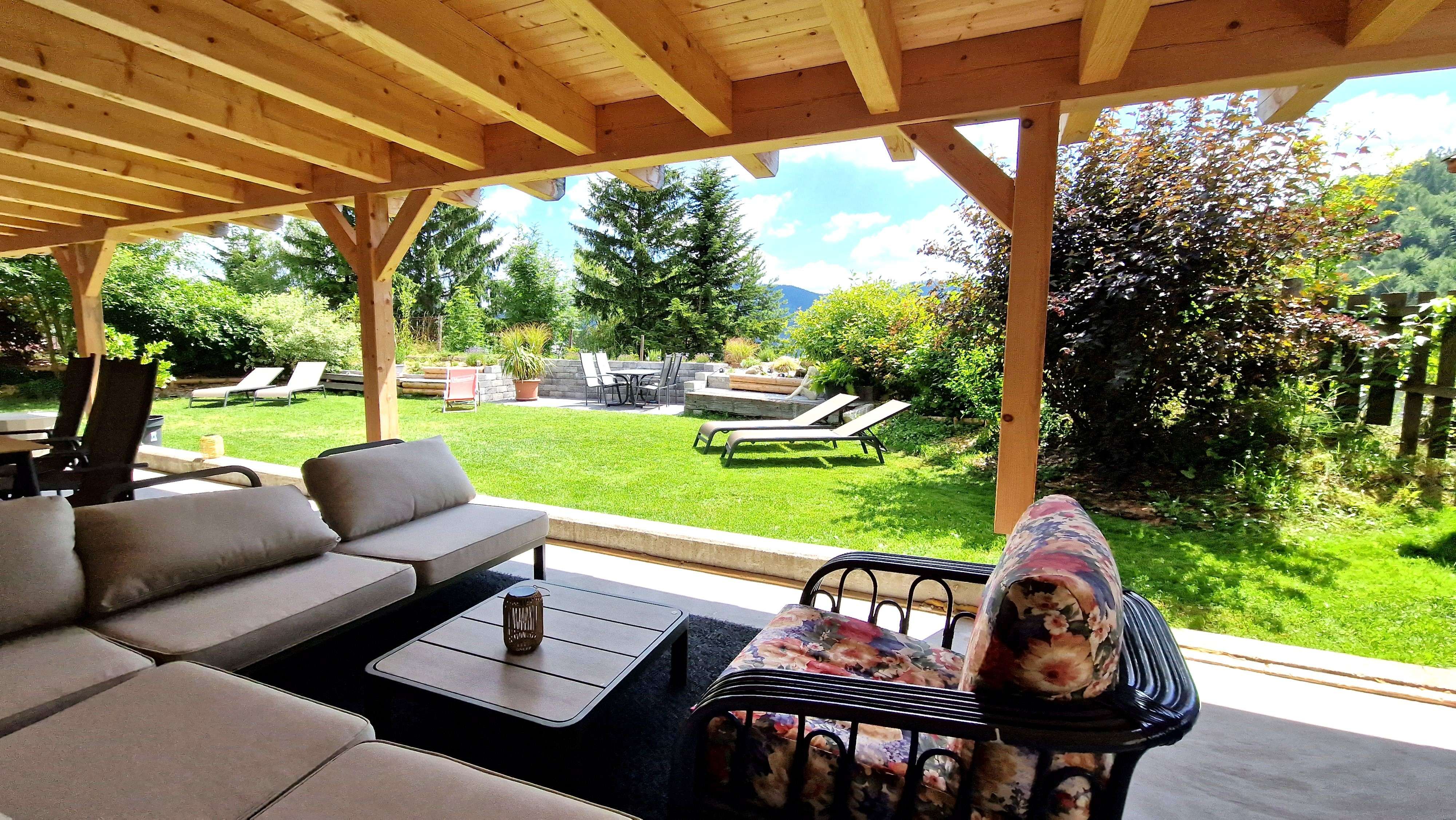 Überdachte Terrasse mit Sitzmöbeln und Blick auf einen Garten mit Liegestühlen und Bäumen.