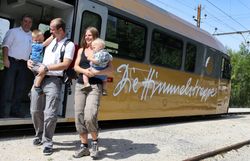 Eine fröhliche Familie steigt aus der bunten Bahn und genießt den warmen Sommertag. Die Kinder lachen und tragen ihre kleinen Rucksäcke, während die Eltern die malerische Umgebung bewundern. Hier beginnt ein unvergessliches Abenteuer in den Bergen.
