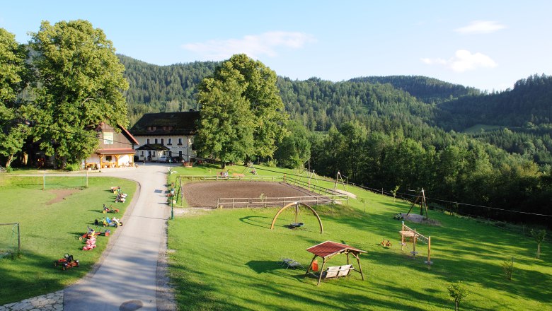 Ein Bauernhof in einer gr&uuml;nen, h&uuml;geligen Landschaft mit Spielplatz und Kettcars auf einer Wiese.
