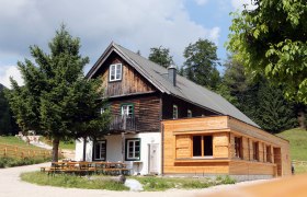 Ein traditionelles Holzhaus mit Anbau in einer l&auml;ndlichen Umgebung, umgeben von B&auml;umen und Wiesen.