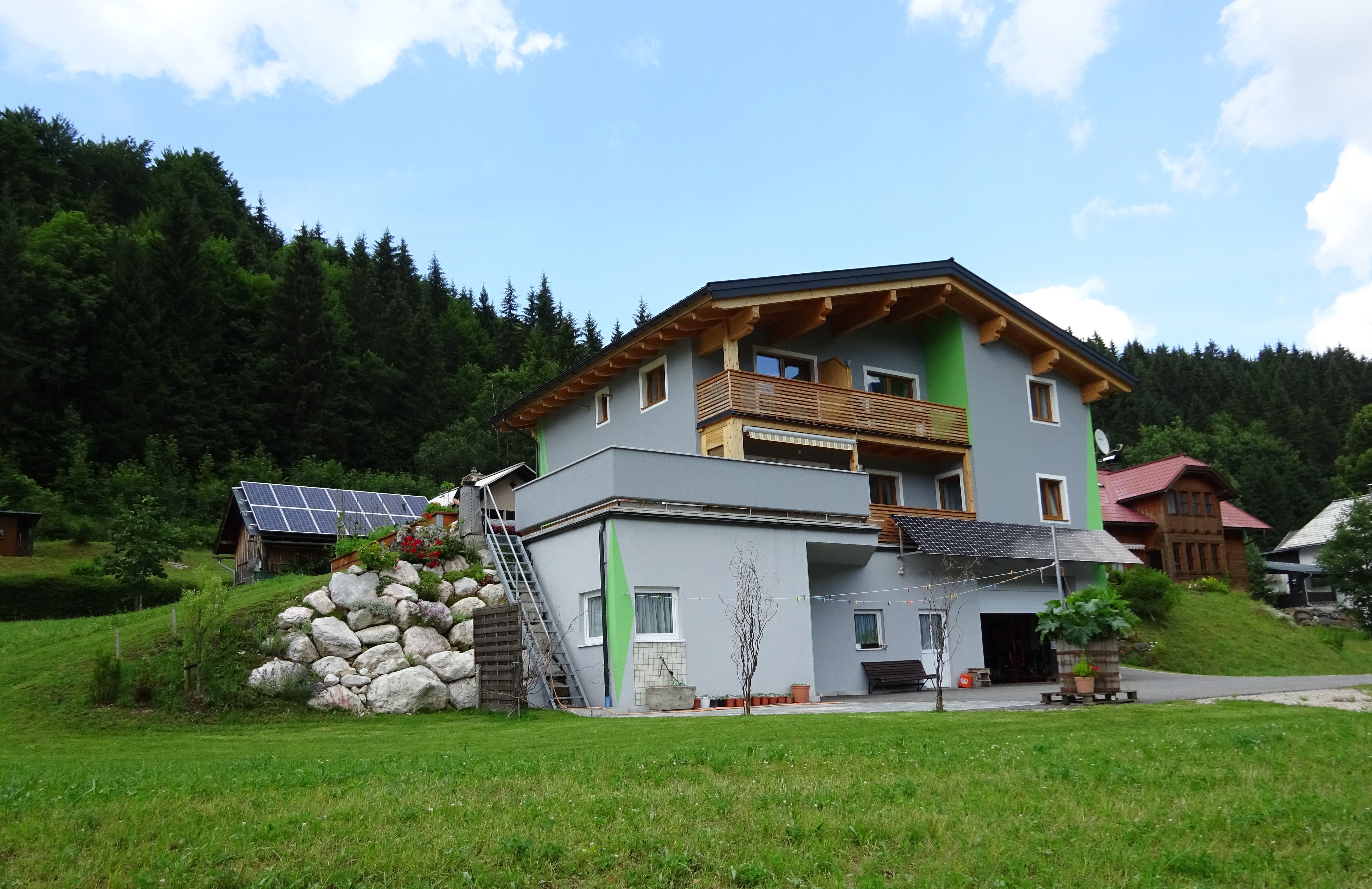 A modern house with wooden cladding and solar panels in a green, wooded setting.