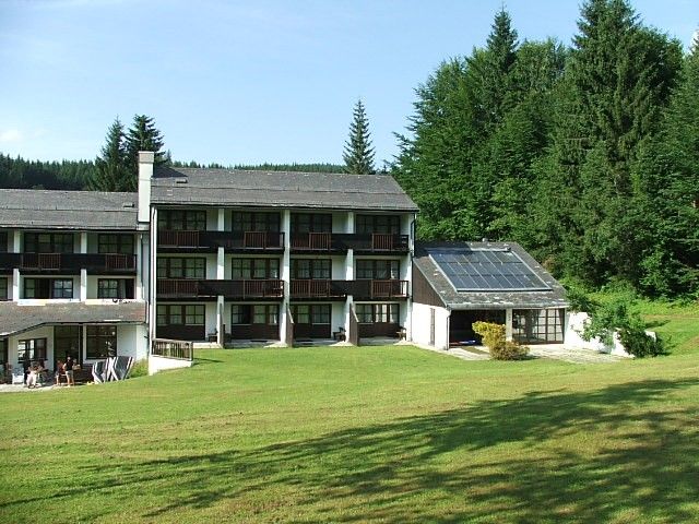 Hotelgebäude mit Solarpanelen und grüner Wiese im Vordergrund, umgeben von Wald.