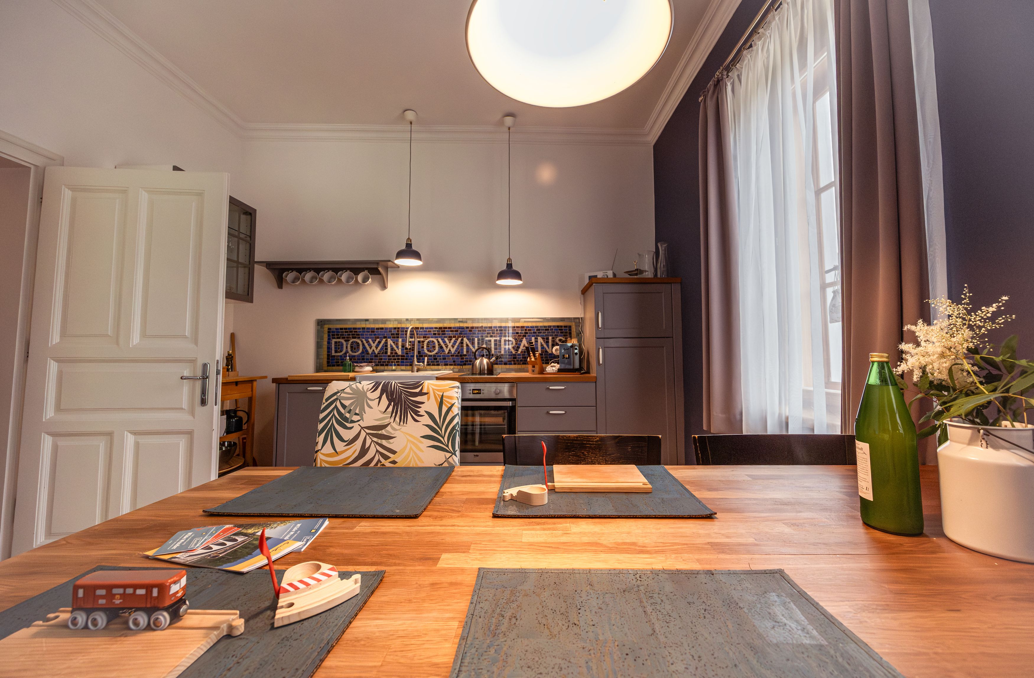 Kitchen with wooden table, toy train, bottle and flowers on the table, modern lamps and kitchen cabinets in the background.