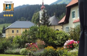 Ein malerischer Garten mit bunten Blumen vor traditionellen H&auml;usern und einer Kirche in einer bergigen Landschaft.
