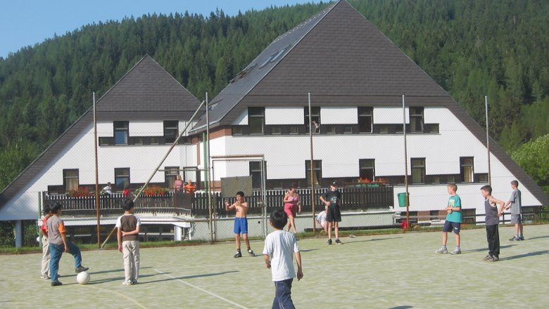 Kinder spielen Fu&szlig;ball auf einem Sportplatz vor einem Hotel in einer bewaldeten Gegend.