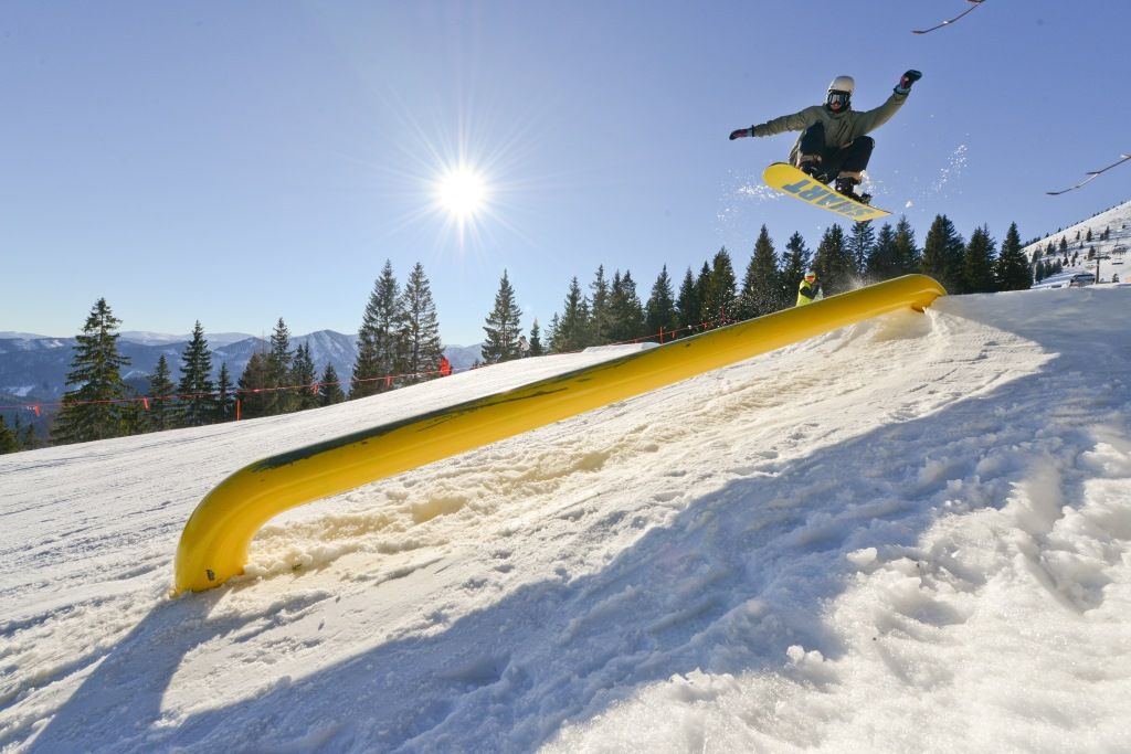 Snowboarder springt über ein gelbes Hindernis im Snowpark Gemeindealpe bei Sonnenschein.
