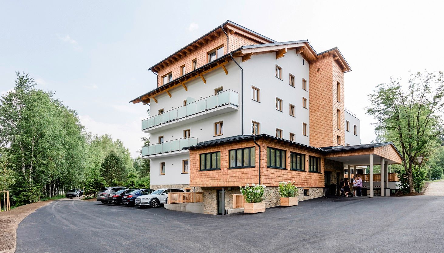 A modern hotel building with wooden cladding and balconies, surrounded by trees and a parking lot with cars.