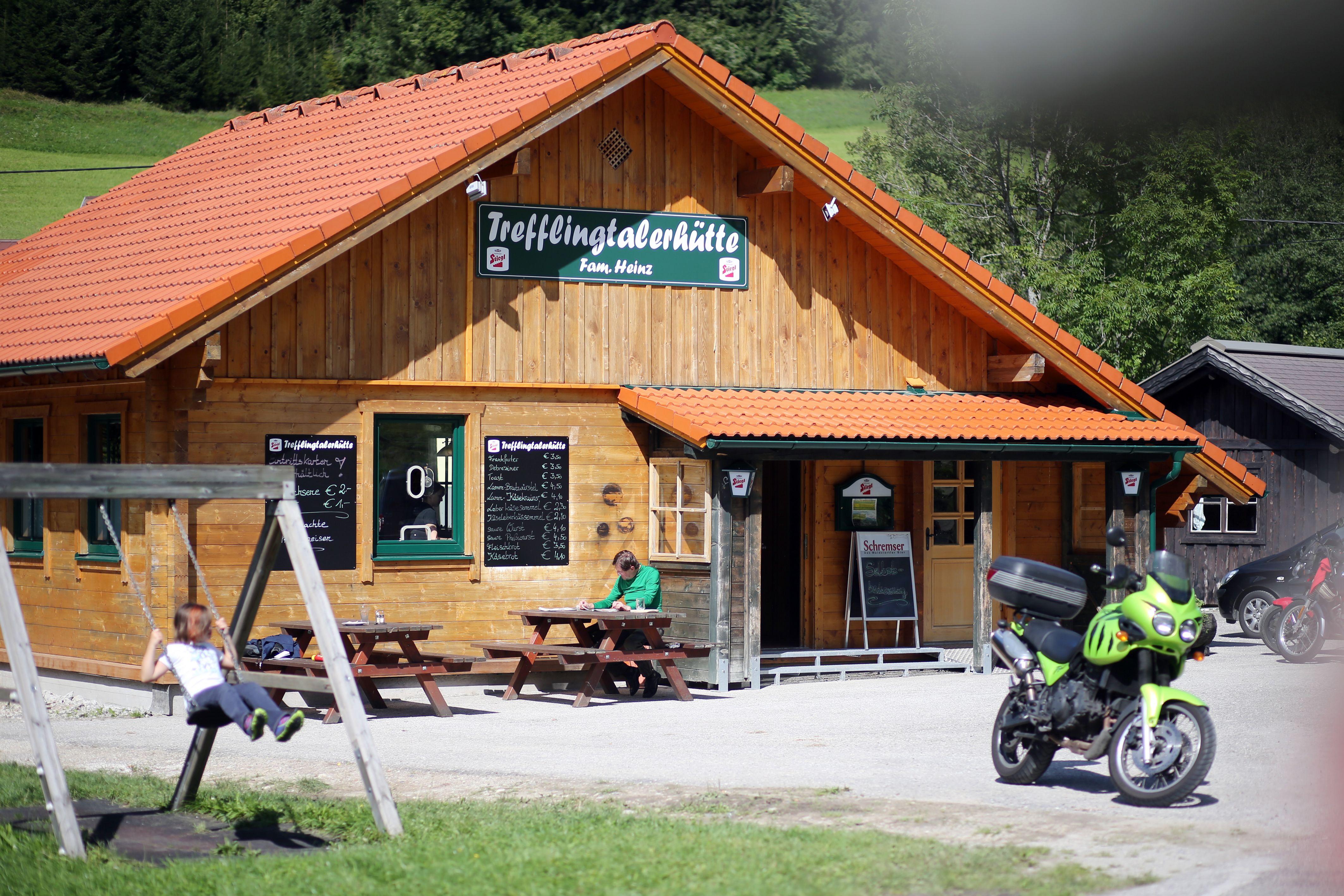 Eine Holzhütte mit dem Schild 'Trefflingtalerhütte', davor eine Schaukel, ein Motorrad und ein Mensch an einem Tisch.