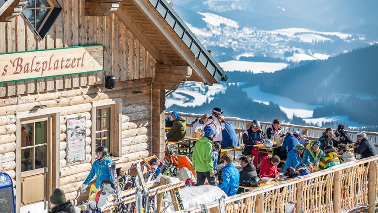 Menschen auf einer sonnigen Terrasse vor einer Berghütte mit Blick auf verschneite Berge.