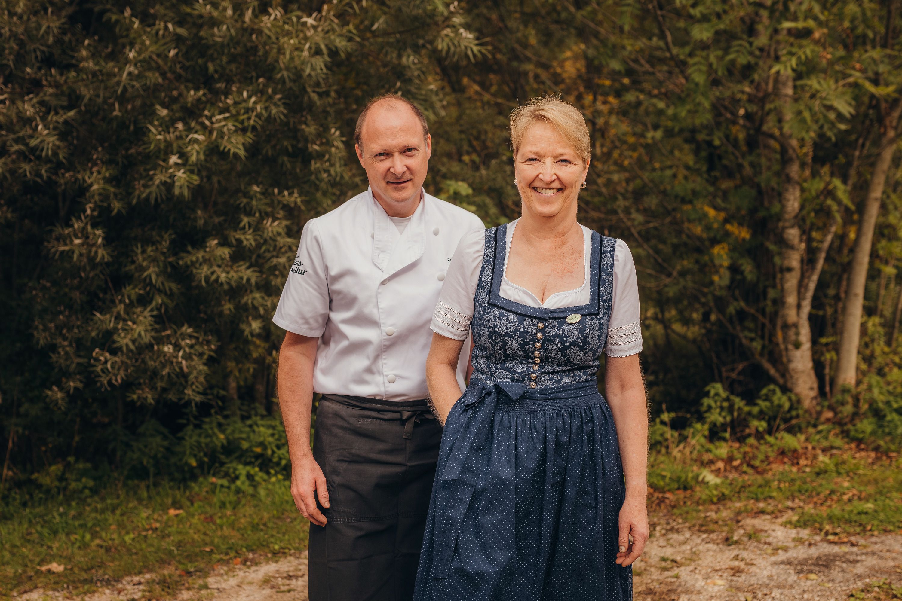 Ein Mann in Kochuniform und eine Frau in traditioneller Tracht stehen lächelnd vor einem Wald.