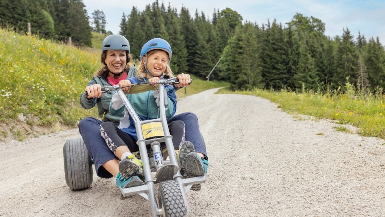 Two people ride a mountain cart on a gravel road surrounded by meadows and forest.