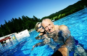 Ein älteres Paar schwimmt lächelnd in einem Freibad mit Wald im Hintergrund.