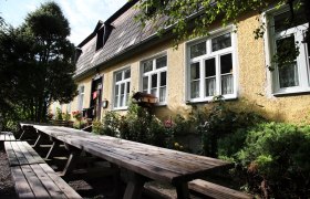 Gelbes Gebäude mit weißen Fenstern und langen Holzbänken im Vordergrund.