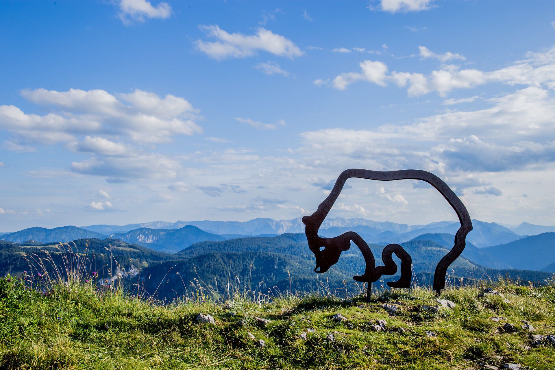 A majestic view of the rolling hills and impressive mountains stretching out under a bright blue sky. The harmonious combination of nature and art is emphasized by the stylish sculpture of the Ötscher bear, which gives hikers a sense of adventure and freedom.