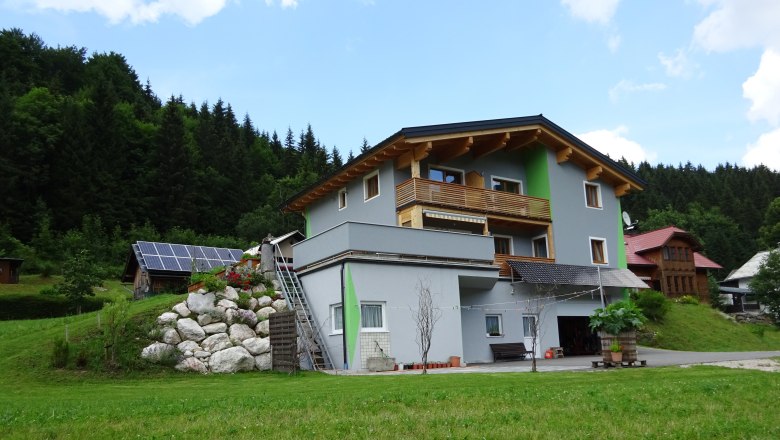 Haus Kölch, © zVg Haus Kölch Ein modernes Haus mit Holzverkleidung und Solarpanelen in einer grünen, bewaldeten Umgebung.