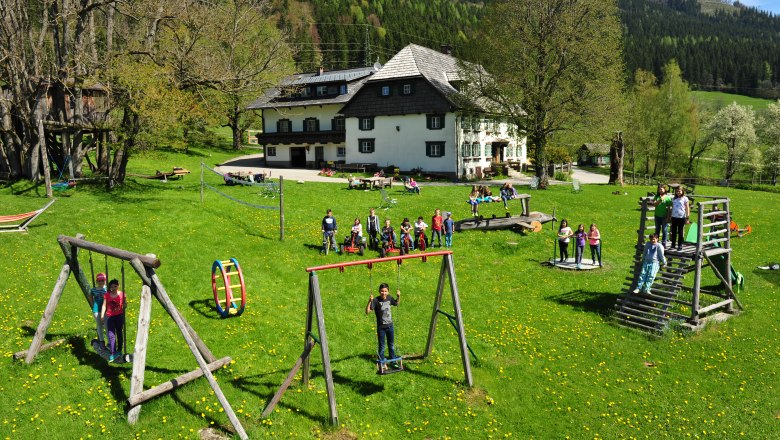 Kinder spielen auf einem Spielplatz vor einem gro&szlig;en Haus in einer gr&uuml;nen Landschaft.