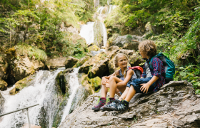 Zwei Kinder sitzen entspannt auf einem gro&szlig;en Stein und genie&szlig;en die erfrischende Atmosph&auml;re des Naturparks. Umgeben von &uuml;ppigem Gr&uuml;n und dem sanften Rauschen des Wassers, strahlt die Szene pure Lebensfreude und Abenteuerlust aus.