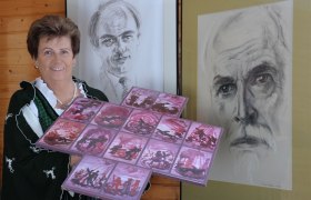 Frau in Tracht h&auml;lt Kunstwerk vor zwei Portr&auml;ts.